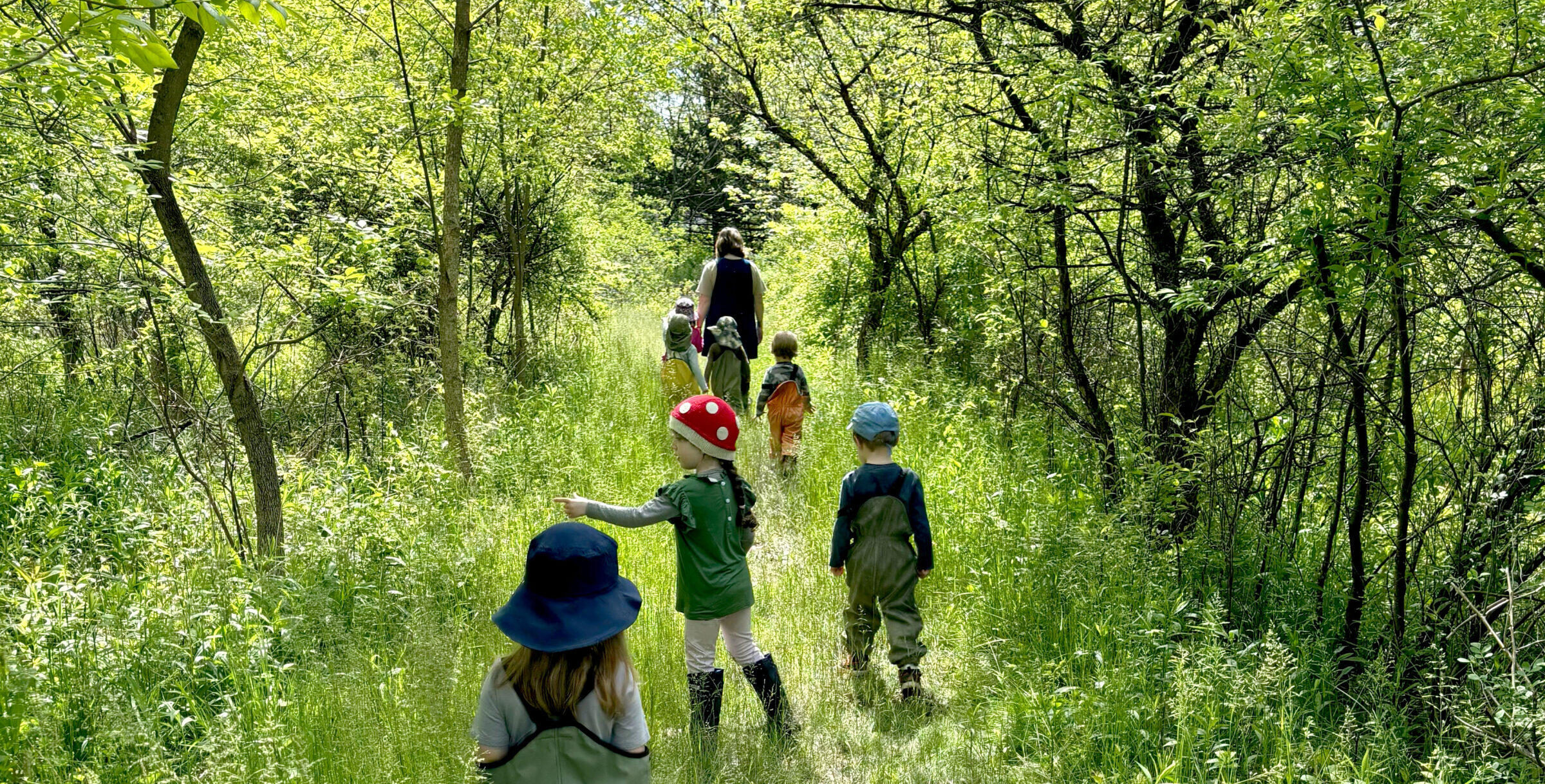 A group of children and their caregiver walking together down a grassy trail in the woods.