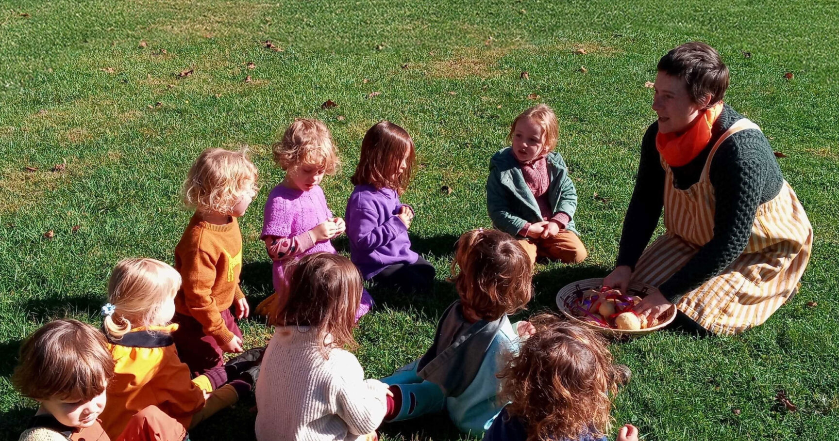 Caitlin sharing a snack with a circle of children seated on the grass.